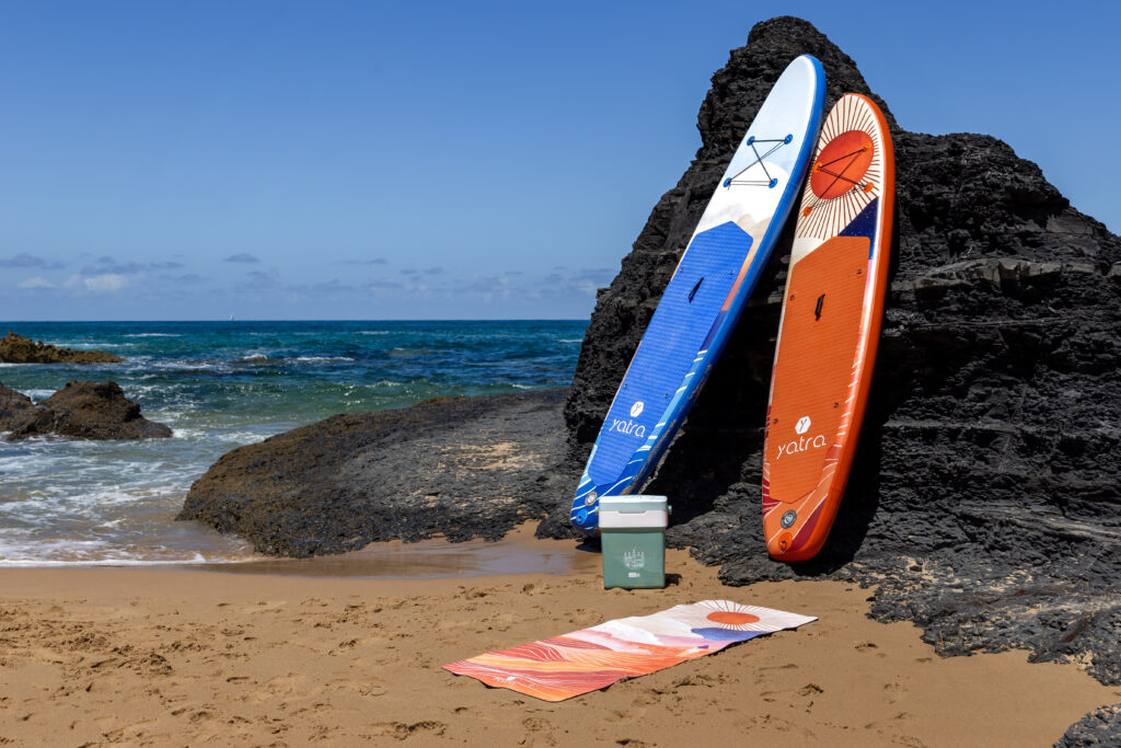 Dwie pompowane deski SUP Yatra Coral Sunrise i Sea Foam ustawione na plaży przy skalistym wybrzeżu